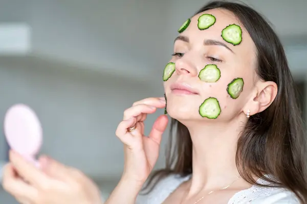 Yüzünde salatalık dilimleri olan bir kadın el aynasına bakar ve sağlıklı cilt bakımından zevk alır.