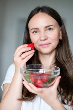 Kadın gülümsüyor ve taze çilek yiyor. Doğal sağlıklı atıştırmalıkların tadını çıkarıyor.