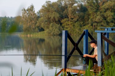 Genç bir çocuk elinde oltayla tahta bir iskelede oturuyor, gölün kenarında sisli bir sabahın tadını çıkarıyor.
