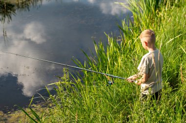Genç çocuk nehir kıyısında doğanın tadını çıkarıyor, yaz tatilinde uzun yeşil çimenlerin üzerinde duruyor.