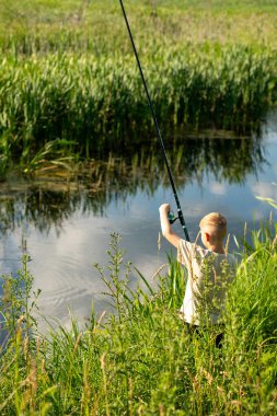 Genç çocuk nehir kıyısında doğanın tadını çıkarıyor, yaz tatilinde uzun yeşil çimenlerin üzerinde duruyor.