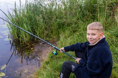 Genç bir çocuk bir yaz günü nehir kenarında balık tutuyor, açık hava ve kırsal atmosferin tadını çıkarıyor.