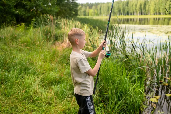 Sarışın çocuk güneşli bir yaz gününde gölün kıyısında balık tutuyor, boş zamanların tadını çıkarıyor, doğada huzurlu bir çocukluk geçiriyor.