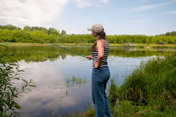Bir oltayla gölün kıyısında duran genç bir kadın, huzurlu bir yaz gününün tadını çıkarıyor, doğada hobi olarak dinleniyor, boş zamanlarında aktiviteler yapıyor.