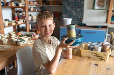 Mutlu çocuk kil bardağını gururla tutuyor, çömlek kıskaçlarında yaratıcı başarının tadını çıkarıyor.