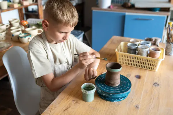 Sarışın çocuk, el yapımı kilden bardakları dikkatlice boyuyor. Rahat ev çömlekçiliğinde yaratıcı çocuk aktiviteleri yapıyor.