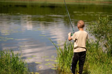 Mutlu sarışın çocuk elinde oltayla göl kıyısında duruyor, parlak bir yaz gününde balık tutuyor, açık hava aktivitelerinin keyfini çıkarıyor, huzurlu doğa ve çocukluk özgürlüğünün tadını çıkarıyor.
