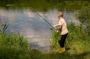 Mutlu sarışın çocuk elinde oltayla göl kıyısında duruyor, parlak bir yaz gününde balık tutuyor, açık hava aktivitelerinin keyfini çıkarıyor, huzurlu doğa ve çocukluk özgürlüğünün tadını çıkarıyor.