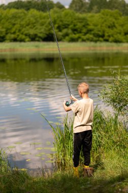 Mutlu sarışın çocuk elinde oltayla göl kıyısında duruyor, parlak bir yaz gününde balık tutuyor, açık hava aktivitelerinin keyfini çıkarıyor, huzurlu doğa ve çocukluk özgürlüğünün tadını çıkarıyor.