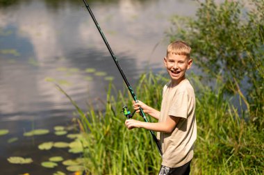 Güneşli bir yaz gününde balık tutarken gülümseyen neşeli sarışın çocuk, nehir kenarında olta tutuyor, doğa eğlencesinin ve çocukluk macerasının tadını çıkarıyor.