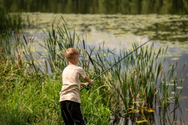 Mutlu sarışın çocuk elinde oltayla göl kıyısında duruyor, parlak bir yaz gününde balık tutuyor, açık hava aktivitelerinin keyfini çıkarıyor, huzurlu doğa ve çocukluk özgürlüğünün tadını çıkarıyor.