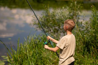 Mutlu sarışın çocuk elinde oltayla göl kıyısında duruyor, parlak bir yaz gününde balık tutuyor, açık hava aktivitelerinin keyfini çıkarıyor, huzurlu doğa ve çocukluk özgürlüğünün tadını çıkarıyor.