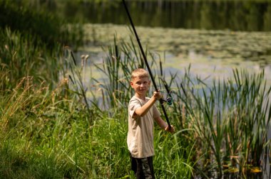 Güneşli bir yaz gününde balık tutarken gülümseyen neşeli sarışın çocuk, nehir kenarında olta tutuyor, doğa eğlencesinin ve çocukluk macerasının tadını çıkarıyor.
