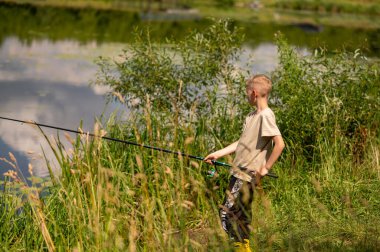 Mutlu sarışın çocuk elinde oltayla göl kıyısında duruyor, parlak bir yaz gününde balık tutuyor, açık hava aktivitelerinin keyfini çıkarıyor, huzurlu doğa ve çocukluk özgürlüğünün tadını çıkarıyor.