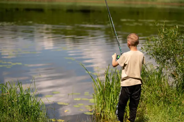 Mutlu sarışın çocuk elinde oltayla göl kıyısında duruyor, parlak bir yaz gününde balık tutuyor, açık hava aktivitelerinin keyfini çıkarıyor, huzurlu doğa ve çocukluk özgürlüğünün tadını çıkarıyor.