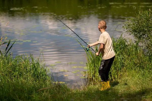 Mutlu sarışın çocuk elinde oltayla göl kıyısında duruyor, parlak bir yaz gününde balık tutuyor, açık hava aktivitelerinin keyfini çıkarıyor, huzurlu doğa ve çocukluk özgürlüğünün tadını çıkarıyor.