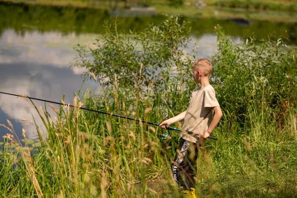 Mutlu sarışın çocuk elinde oltayla göl kıyısında duruyor, parlak bir yaz gününde balık tutuyor, açık hava aktivitelerinin keyfini çıkarıyor, huzurlu doğa ve çocukluk özgürlüğünün tadını çıkarıyor.
