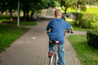 Sıcak ve güneşli bir günde park yolunda bisiklet süren sarışın gencin arka görüntüsü özgürlük, doğa ve aktif yaz yaşam tarzının tadını çıkarıyor.