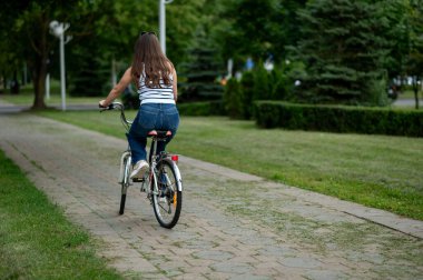 Parlak bir yaz parkında, ağaçlık bir patika boyunca bisiklet süren orta yaşlı bir kadının arka görüntüsü, barışçıl açık hava aktivitesinin ve temiz havanın tadını çıkarıyor.