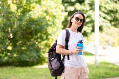 Aktif bir kadın, sıcak ve güneşli bir günde parkta sırt çantası ve spor şişesiyle susuzluk ve temiz havanın tadını çıkarıyor.