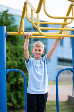 Odaklanmış sarışın çocuk açık hava antrenmanı sırasında yatay barlara takılır, kuvvet kazanır ve şehir parkında eğlenir.