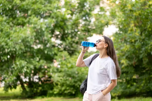 Sırt çantası takan aktif bir kadın parkta durup su, doğa ve açık havada güneşli bir günün tadını çıkarırken spor şişesinden içki içiyor.