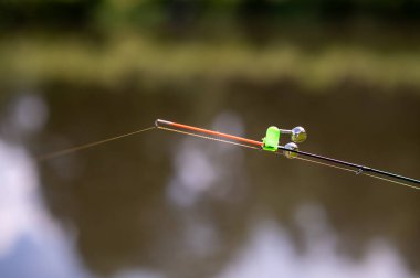Çan ısırığı alarmı takılı oltanın ucu yakın plan, sakin su yüzeyinin bulanık arkaplanı.