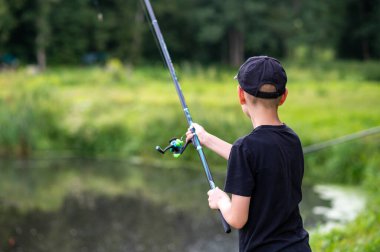 Genç bir çocuk, güneşli yaz güneşinin altında sakin bir göle olta atıyor. Açık hava keyfi ve doğanın tadını çıkarıyor.