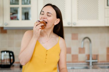 Keyifli bir kadın sıcak bir mutfakta tatlı bir hamur tatlısı yiyor. Zevk ve hoşgörüyü ifade ediyor.
