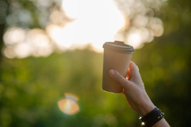 Bir kadın elinde sıcak bir içecekle kağıttan bir bardak tutar, günbatımının altın ışığıyla yıkanmış bir ormanla, sonbaharın sıcaklığını ve rahatlığını anımsatır..