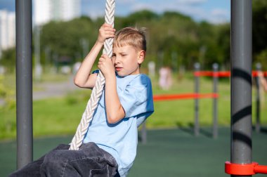 Genç çocuk kendini halatla yukarı çekiyor. Spor sahası, eğitim anlayışı, güç, dayanıklılık ve aktif yaşam tarzı için çabalıyor.