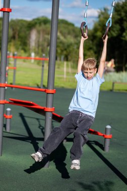 Sarışın genç çocuk açık havada jimnastik ringlerinde sallanıyor, eğlence, güç, çeviklik ve aktif yaşam tarzı kavramı