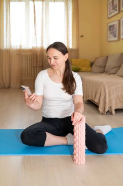  Kadın yoga minderinde köpükle oturuyor, telefona bakıyor, egzersiz sonrası rahatlama ve sağlık izleme konseptine bakıyor.