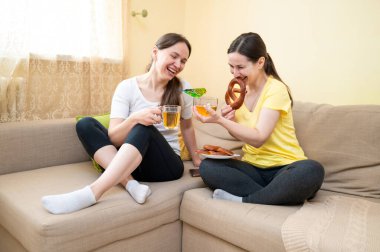 İki arkadaş kanepede keklerle gülüyor ve çay içiyor, ve yeşil muhabbet kuşu fincanın üzerine düşüyor, ev konforu ve neşe anı.