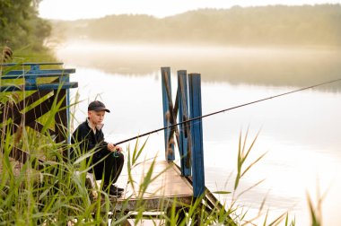 Küçük çocuk, sisli bir gölün kıyısındaki ahşap iskelede tek başına balık tutmayı öğreniyor.