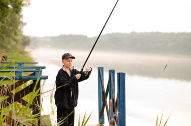 Küçük çocuk, sisli bir gölün kıyısındaki ahşap iskelede tek başına balık tutmayı öğreniyor.