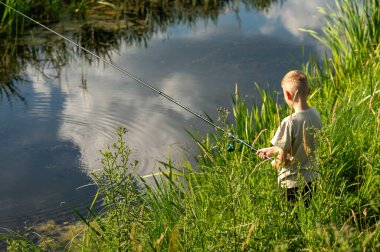 Genç çocuk nehir kıyısında doğanın tadını çıkarıyor, yaz tatilinde uzun yeşil çimenlerin üzerinde duruyor.