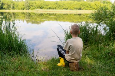 Sarışın çocuk göl kıyısında oltayla balık tutuyor, doğada huzurlu bir yaz gününün tadını çıkarıyor, açık havada dinleniyor, çocukluk tatili yapıyor.