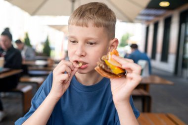 Aç sarışın genç bir çocuk güneşli bir kafede sulu bir burgerin tadını çıkarıyor, yaz yemeği sırasında iştahını ve neşesini açık hava setiyle gösteriyor.