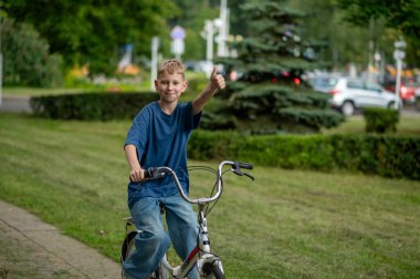 Sarışın genç çocuk bisikletini ağaçlarla kaplı bir park yolunda sürüyor, sıcak havanın, rahatlamanın ve doğada neşeli aktivitelerin tadını çıkarıyor.
