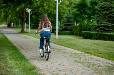 Parlak bir yaz parkında, ağaçlık bir patika boyunca bisiklet süren orta yaşlı bir kadının arka görüntüsü, barışçıl açık hava aktivitesinin ve temiz havanın tadını çıkarıyor.