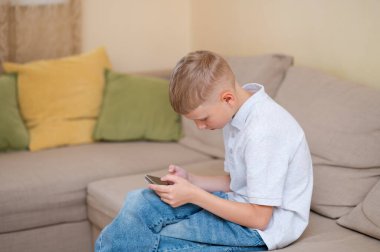 Genç çocuk bir kanepede oturuyor, akıllı telefonuna dalmış, modern alet bağımlılığını ve gençler arasındaki aşırı ekran zamanını resmediyor.