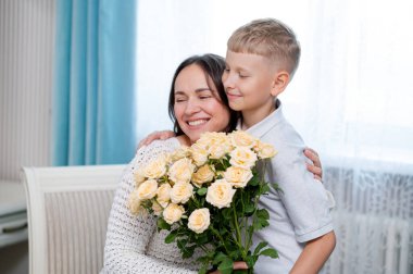 Ergen çocuk sıcak bir odada anneler gününü sevgi ve minnettarlıkla kutlayan sarı gülleri sunarken annesine sıcak bir şekilde sarılıyor.