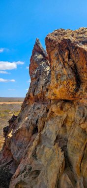 Açık mavi gökyüzünün altında pürüzlü dokuları olan yüksek kaya oluşumu. Güneş ışığı, engin manzaraya karşı toprak tonlarını vurgular, ihtişam ve zamansızlık hissini taşır.
