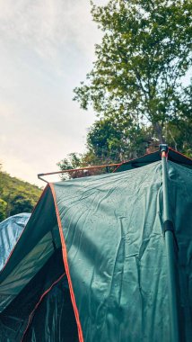 Yüzeyinde su damlacıkları olan yeşil bir çadır, doğal bir dağ ve ağaç zeminine karşı duruyor. Bulutlu gökyüzü serin ve sakin bir atmosfer ekler. Macera duyguları ve doğa ile bağlantı hissi uyandırır..