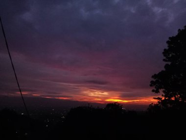 Görüntü, bir şehir manzarası üzerinde nefes kesici bir gün batımı ya da gün doğumu görüntüsü yakalar, gökyüzü ufukta koyu mor ve koyu mavi renkten parlak turuncu ve kırmızı renge dönüşür.