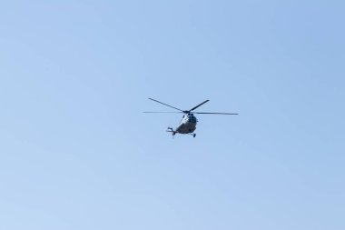 Bir helikopter açık gökyüzünde süzülüyor, açık mavi bir zemin üzerine çerçevelenmiş, özgürlük ve macera hissi veriyor..