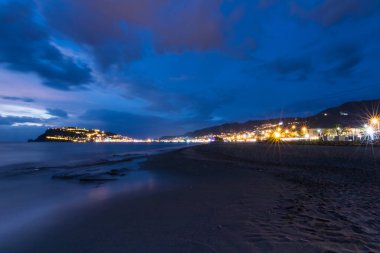 Karanlık çöktüğünde deniz kenarı şehri aydınlandı. Kasvetli bir kumsaldan çekiliyor. Alacakaranlık atmosferi ve manzaralı bir ufku var..