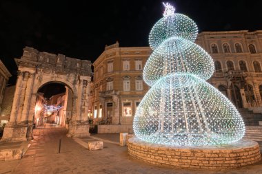 Kadim bir taş kemer ve yakınlardaki yapılar gece şenlikli süslemelerle parlayarak büyülü bir tatil atmosferi ve çarpıcı mimari tezatlıklar yaratıyor. Altın kapı, Pula..