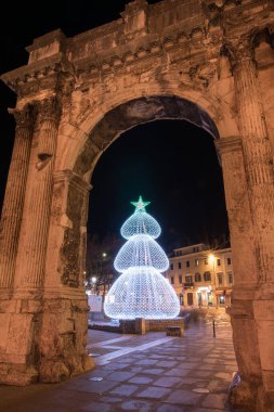 An ancient stone arch and nearby structures brightly light up with festive decorations at night, creating a magical holiday atmosphere and stunning architectural contrasts. Golden door, Pula. clipart